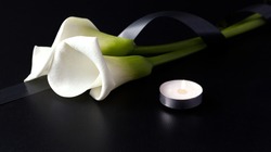 White Zantedesia with mourning ribbon and burning candles on a black background. Concept of sorrow and death