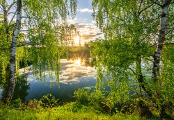 Scenic view at beautiful spring sunset on a shiny lake with green branches, birch trees, bushes, grass, golden sun rays, calm water ,deep blue cloudy sky and forest on a background, spring landscape