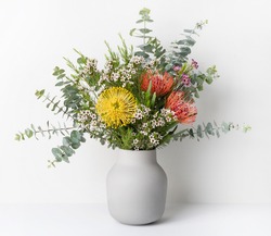 A beautiful floral arrangement of Australian native red and yellow Waratah flowers,  eucalyptus leaves and wax flowers in a stylish grey vase, with a white background.