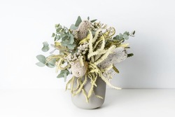 Beautiful flower arrangement of Australian native dried banksia, eucalyptus leaves and delicate white flowers, in a grey vase on a table with a white background.