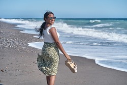 Young African American woman walking along the shore of the beach with sandals in hand