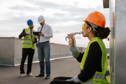 On construction site beautiful smiling African lady engineer drinking some water from bottle on the rooftop of building she wearing safety helmet and yellow safety glasses