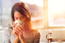 Woman drinking coffee at home with sunrise streaming in through window and creating flare into the lens.