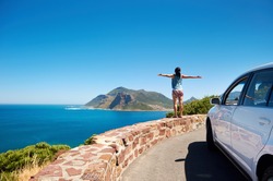 carefree tourist stands on chapmans peak drive with arms outstretched in freedom girl pose with rental car