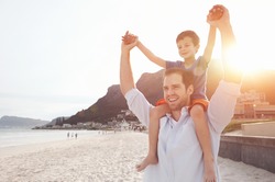 Son on fathers shoulders at the beach having fun at sunset together
