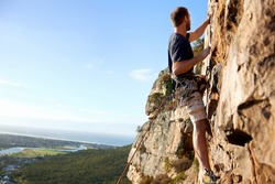 A male rockclimber climbing up a steep mountain attached to a rope and harness looking at the view