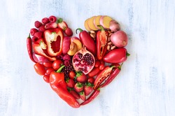 Collection of fresh red vegetables and fruits arranged in a heart shape on white rustic background strawberry raspberry pomegranate peppers capsicum chilli potato beans legumes overhead flat lay