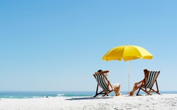 Beach summer couple on island vacation holiday relax in the sun on their deck chairs under a yellow umbrella. Idyllic travel background.