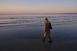 Stylish girl walks at sunset along the beach of the sea or ocean on a cool day. Travel concept. Walk on the sea or ocean. Girl in travel. Amazing scenic outdoors view. Beautiful sunset on the seashore