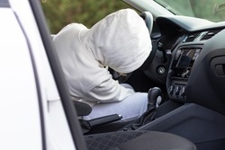 A man in a white sweater with a hood sits at the wheel of a car and tries to start it in order to steal it. Selective focus. Close-up