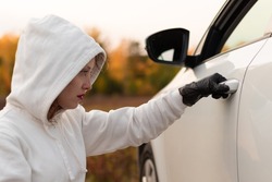 A beautiful young woman in a white sweater with a hood and black gloves opens the car door to steal it. Selective focus. Close-up. Portrait