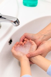 Soap foam in the shape of a heart in children's hands over the washbasin and mother's hands in a bright bathroom. Selective focus. Close-up