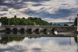 Old arched bridge crossing the still Vartry River in Wicklow town. Historical stone bridge with arches across the calm water of a river in the countryside village of Wicklow shows a typical Irish road