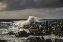 Rough ocean waves smashing on hexagonal rocks at the giant 's causeway on a murky day. Hexagon - shaped stones with a sea wave hitting the cliff convey a strong impact or major, big effect concept