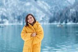 winter travel across Europe. portrait of a beautiful young woman in a yellow jacket on the shore of an alpine lake in winter. view of the alpine lake with snow.