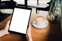 A woman hands are holding a mockup of black tablet with blank white screen   in the cafe with a white cup of latte coffee. sitting on the wood table, clipping path included