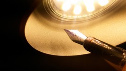An old shiny fountain pen featuring a golden metal nib illuminated by the warm yellow light of a LED lamp in the conical form of a cupola ; symbolic image for inspiration in literacy or revelation
