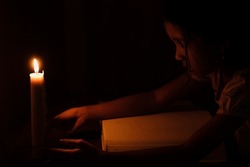 Little Asian girl is reading in a dark room lit only by candlelight. The concept of children's education in a village without electricity.