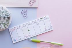 7 days week planner with pen, paperclips, phone and succulent on a pink and blue background.