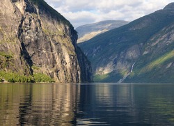Mountains Reflecting In The Calm Water Of Geirangerfjord With The Friaren Falls In The Background On A Sunny Summer Day With A Clear Blue Sky And A Few Clouds