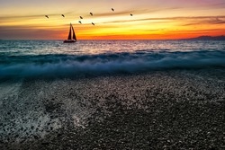 A Sailboat Is Sailing Along The Ocean Against A Colorful Sunset Sky With Birds Flying Overhead