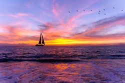 Ocean sunset sailboat silhouette is sailboat sailing along the ocean water with a colorful vivid sunset sky and silhouettes of birds flying in the background..