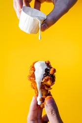 Hand holding a chicken wing with buffalo and ranch dressing on a yellow background.
