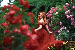 portrait of a beautiful young girl with red hair and a red dress in a garden of roses 