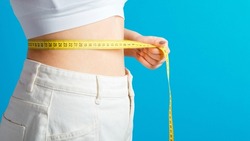 Slim woman measures her waistline with measuring tape. Healthy body shaping weight loss concept. Slim waist small belly in big white denim pants isolated over blue color background. Long web banner