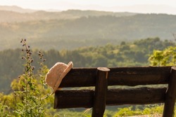 Hat on wooden rustic bench against background of nature mountains and atmospheric epic forest valley. Peaceful, quiet, serene summer evening in sunset light while travel, vacation or weekend getaway