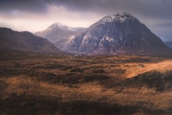 Dramatic golden light on the dark, moody, mountain landscape of Buachaille Etive Mor with a distant Blackrock Cottage at Glencoe in the Scottish Highlands, Scotland.