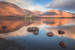 Scottish Highlands landscape of the historic ruins of Kilchurn Castle reflected on a calm, peaceful Loch Awe with sunset or sunrise golden light.
