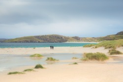 A distant couple take a leisurely stroll along the white sand beach and grassy dunes at Reef Beach on the Isle of Lewis and Harris in the Outer Hebrides of Scotland, UK.