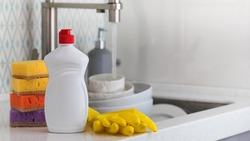 A white bottle with dishwashing gel, sponges and rubber gloves on the background of a sink with dirty dishes. Copy space
