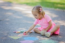 Concentrated toddler girl sitting and drawing shapes on asphalt. Child holding chalk. Street art, kids education. Natural light. Selective focus.