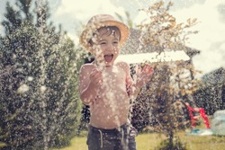 Cute little boy in straw hat is laughing and having fun running under water spraying hose. Image with selective focus and toning