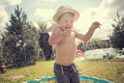 Cute little boy in straw hat is laughing and having fun under water spraying hose. Image with selective focus and toning