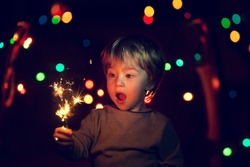 Cute little boy with a funny face holding sparklers. There is a background with multi colored lights. Image with selective focus and toning