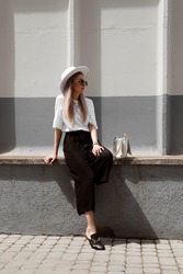 portrait of a blonde woman in a stylish summer fashion look, girl sitting in a white oversize t-shirt, black linen pants, mules, glasses and white hat, full length model