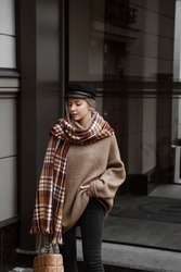 portrait of beautiful blonde girl in trendy outlook, model dressed in black flared jeans with slits, brown oversize sweater, cap, loafers, scarf, bag in hand, stylish autumn fashion look, lifestyle