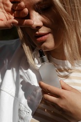 portrait of beautiful blonde girl with plump lips, model holds matte jar of perfume or cosmetics in hand near the face, sun glare and shadows on face, hiding from the sun, advertising, close up