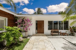 Beautiful entrance of house with cement and stone floors, tropical plants, windows, outdoor chair and table, white walls, blue sky located in Pinecrest, Miami-Dade