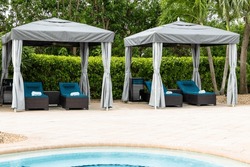 Point of view from lounge chairs with rolled towel in gazebo with fan, swimming pool, surrounded by greenery and tropical plants, summer weather, blue sky