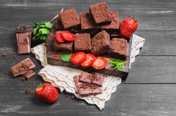Pieces of chopped chocolate cake brownie with nuts, chocolate bars, leaves and sprigs of mint, strawberry berries on a cutting board on a black wooden background
