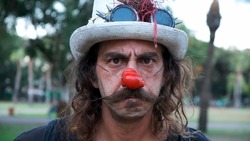 Sad or angry clown portrait. Clown with a mustache and a red nose outdoors in a park. looking to camera with a mad face  