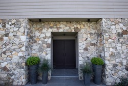 Modern architecture. Residential facade design. Closeup view of new house iron front door, stonewall and decorative plant Buxus sempervirens, also known as boxwood bush, growing in pots. 
