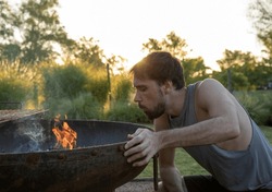 Barbecue in the garden at sunset. Portrait of a young man blowing the fire in the grill. The flames and smoke coming out. 