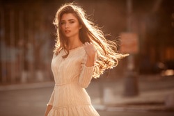 Beautiful young lady with long healthy hair and cute dress walking on the street. Hot summer evening. Sunset. Lifestyle concept.