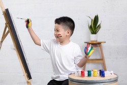 Cute happy little Asian boy painting with water color on canvas standing in white room at home, creative young artist at work. Kids paint. Children draw.