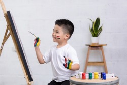 Cute happy little Asian boy painting with water color on canvas standing in white room at home, creative young artist at work. Kids paint. Children draw.